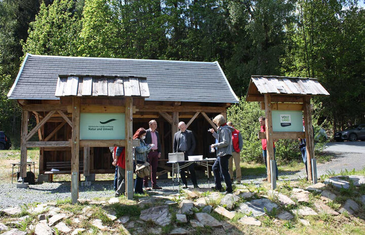 Vor einem Holzunterstand am Waldrand stehen mehrere Erwachsene um einen Tisch. Eine der Personen hält einen kleinen Gegenstand hoch und erklärt etwas. - Link: Erlebnispfad Flussperlmuschel